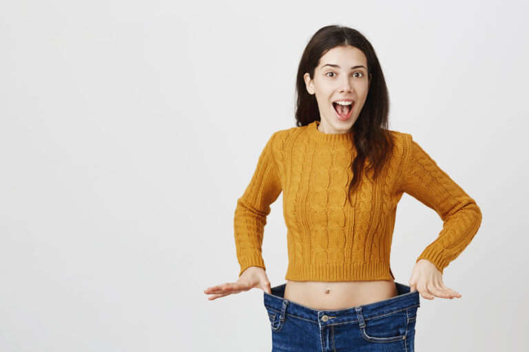 Astonished and happy young lady being impressed and excited because of losing weight, showing empty space in jeans by stretching it, standing over gray background